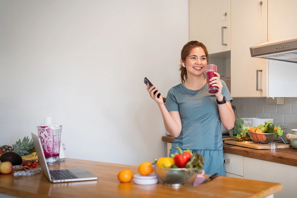 Maya Thompson smiling while working on recipes for Bring Dish on her laptop, holding a smoothie and her phone.