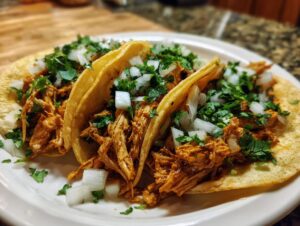 Close-up of three soft corn tortillas filled with shredded chicken tacos, topped with diced white onion and fresh cilantro.
