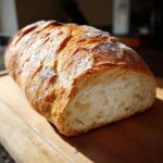 A freshly baked loaf of homemade french bread, sliced open to show the airy crumb, resting on a wooden cutting board.