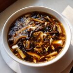 A close-up of a white bowl filled with rich, dark broth of hot and sour soup recipe, showing tofu, mushrooms, and egg ribbons.
