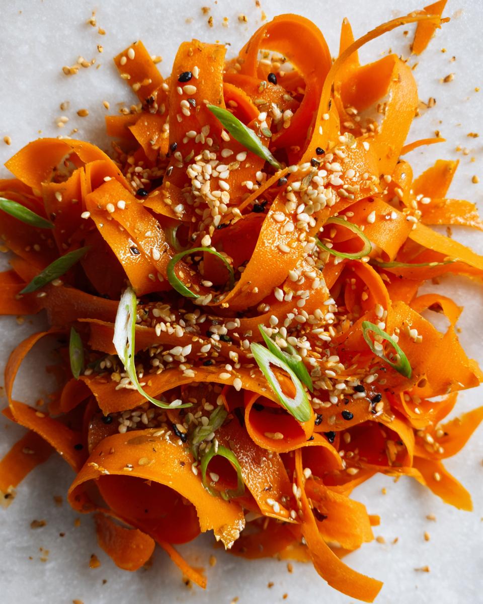Close-up of a vibrant raw carrot salad made with shaved carrot ribbons, topped heavily with sesame seeds and sliced green onions.