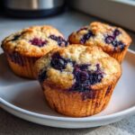 Three golden-brown blueberry protein muffins with visible baked blueberries sitting on a white plate.