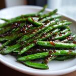 Close-up of bright green, glossy chinese green beans with garlic sauce drizzled over them on a white plate.