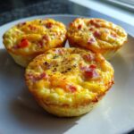 Three golden brown cottage cheese egg cups starbucks style, topped with bacon bits and herbs, sitting on a white plate.