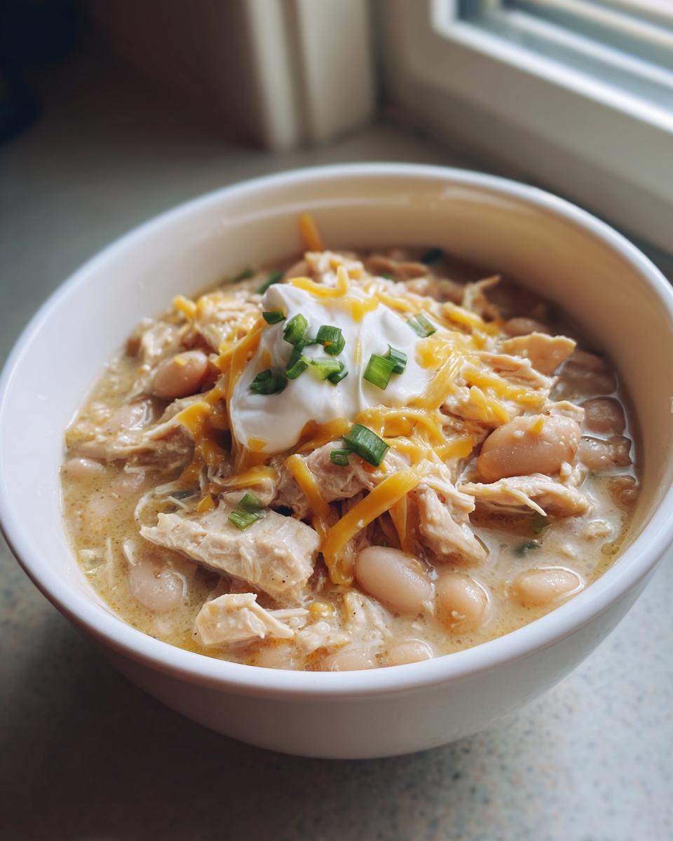 A close-up of a white bowl filled with creamy crockpot white chicken chili, topped with shredded cheese, sour cream, and green onions.