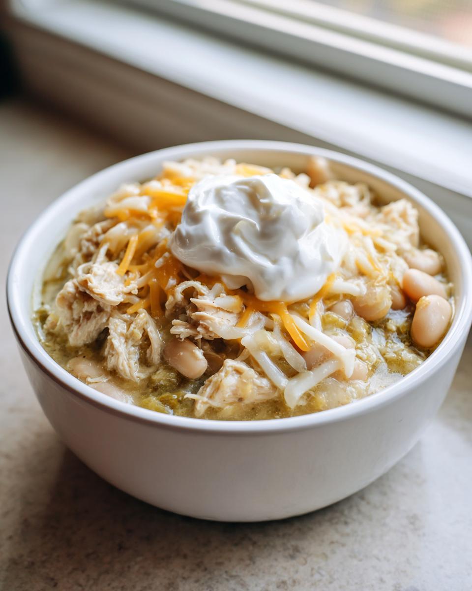 A close-up of a hearty bowl of crockpot white chicken chili topped with shredded cheese, chicken, and a dollop of sour cream.