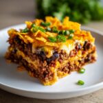 A close-up of a layered slice of Dorito Casserole topped with melted cheese, crushed chips, and green onions.