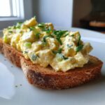 Close-up of creamy egg salad recipe piled high on a slice of toasted whole wheat bread, topped with fresh green chives.