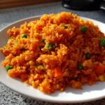 Close-up of a white plate piled high with vibrant orange Spanish rice mixed with peas and carrots.