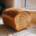 A golden-brown, freshly baked whole wheat bread loaf resting on a wire cooling rack.