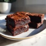 Close-up of fudgy date brownies stacked on a speckled white plate, showing their rich, dark interior.
