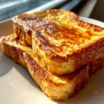 Close-up of two thick slices of golden brown french toast stacked on a white plate, glistening with syrup.