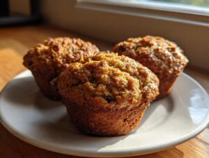 Three freshly baked honey bran muffins with golden, textured tops sitting on a light-colored plate near a window.