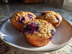 Three freshly baked protein muffins studded with dark purple berries, resting on a light ceramic plate.