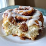 A close-up shot of a single, soft cinnamon roll with rich brown filling and thick white icing, resting on a white plate.