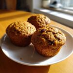 Three freshly baked honey bran muffins, one topped with raisins, resting on a white plate near a sunlit window.