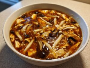 A close-up of a white bowl filled with rich, dark broth containing tofu, mushrooms, and ribbons of egg from the hot and sour soup recipe.