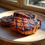 Three perfectly grilled and glazed pork steak recipe cuts resting on a light-colored plate, glistening in the sunlight.