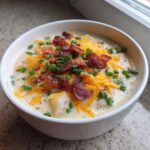 A close-up of a creamy bowl of Loaded Baked Potato Soup topped with shredded cheddar cheese, bacon bits, and fresh chives.