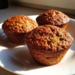 Close-up of three golden-brown, moist bran muffins sitting on a white plate, highlighted by bright sunlight.