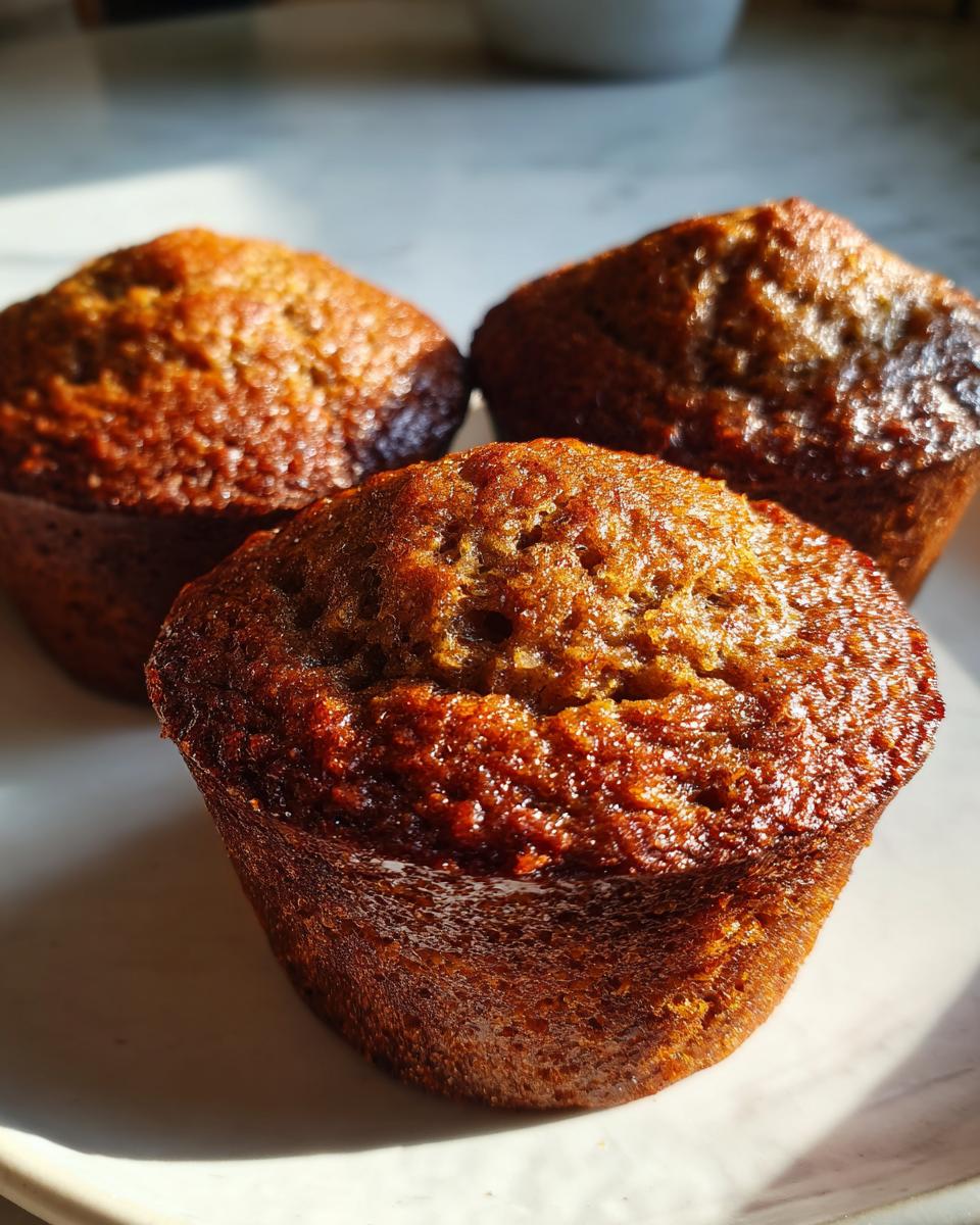 Close-up of three freshly baked, moist bran muffins with a dark, glossy brown top, sitting on a light surface.