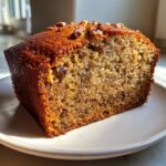 Close-up of a moist slice of sugar free banana bread topped with chopped pecans, served on a white plate.