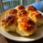 Six freshly baked, golden brown, and glossy dinner rolls arranged on a white plate near a bright window.
