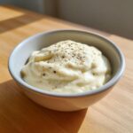 A close-up of thick, creamy homemade alfredo sauce topped with cracked black pepper in a small white bowl.