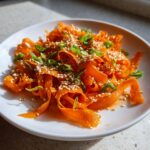 Close-up of a bright orange raw carrot salad made with shaved carrot ribbons, topped with toasted sesame seeds and sliced green onions.