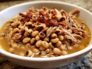 Close-up of a white bowl filled with savory black eyed peas, shredded pork, and crispy bacon pieces.