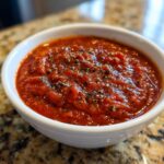 Close-up of a vibrant red, thick pizza sauce seasoned with dried herbs in a small white bowl.