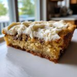 A close-up of a thick slice of chocolate chip cookie cake recipe topped with creamy white frosting.