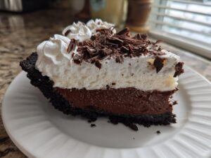 A close-up slice of rich chocolate mud pie with a dark crust, white mousse layer, whipped cream, and chocolate shavings.