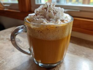 A glass mug filled with a rich, orange pumpkin spice latte topped with whipped cream and a dusting of spice.