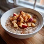 A close-up of a white bowl filled with creamy apple cinnamon oatmeal, topped generously with diced red apples and sprinkled cinnamon.