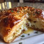 Close-up of baked brie wrapped in golden puff pastry, cut open to show the melted cheese oozing out, garnished with fresh thyme.