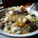 Close-up of a fork lifting a cheesy, stringy bite from a bowl of philly cheese steak soup with beef and green peppers.