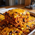 A spoonful of cheesy, orange-tinted mexican rice casserole mixed with black beans and corn being lifted from the baking dish.