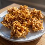 Several golden brown, chewy haystack cookies made with chow mein noodles, displayed on a white plate.