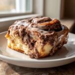 A single, decadent chocolate cinnamon roll covered in thick white icing, sitting on a white plate.