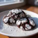 Three rich, dark chocolate crinkle cookies dusted heavily with powdered sugar on a white plate.