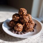 A stack of homemade chocolate protein balls made with oats, served on a white plate with scattered chocolate chips.