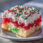 Close-up of a slice of Christmas poke cake showing layers of white cake, green and red gelatin, whipped topping, and festive sprinkles.