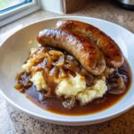 Two perfectly browned sausages served over creamy mashed potatoes, topped with caramelized onions and rich gravy for an amazing bangers and mash.