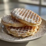A stack of three cream-filled pizzelle cookies dusted heavily with powdered sugar on a light plate.
