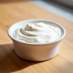 Close-up of thick, creamy homemade garlic sauce served in a small white ramekin on a wooden surface.