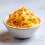 Close-up of thick, bright orange nacho cheese sauce swirled high in a small white bowl.
