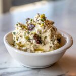 A close-up of a creamy, textured olive dip topped with chopped herbs and whole dark olives, drizzled with olive oil.