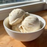 Three scoops of creamy vanilla-colored protein ice cream served in a white bowl on a wooden surface.