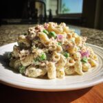 A generous serving of creamy Tuna pasta salad piled high on a white plate, featuring elbow macaroni, green peas, and red onion chunks.
